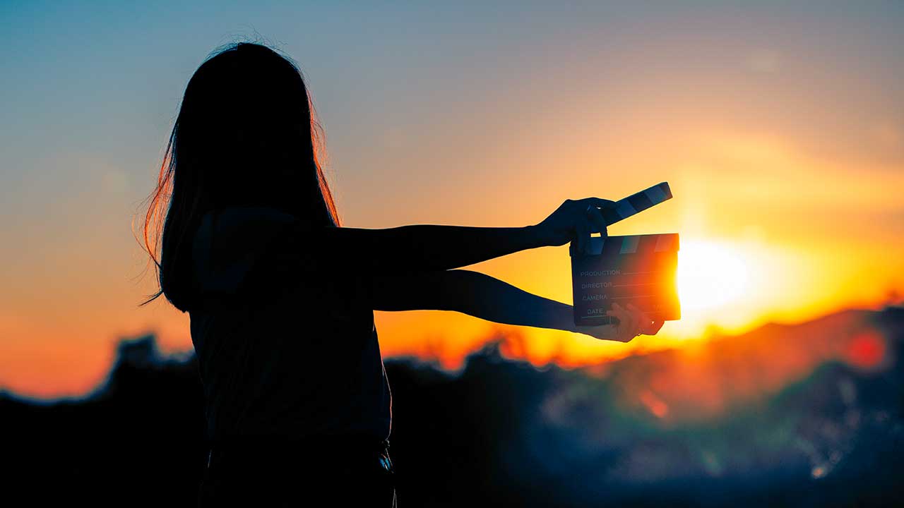 Silhouette einer junge Frau, welche eine Filmklappe vor einen Sonnenaufgang hält