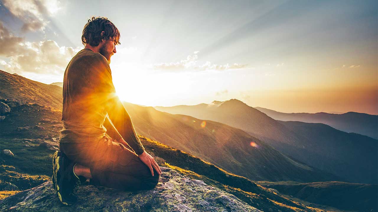 Mann kniet auf einem Fels im Gebirge und wird dabei von Sonnenlicht beschienen