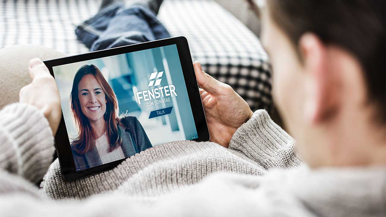 Mann sitzt bequem und hält ein Tablet, auf dem der Vorspann von FENSTER ZUM SONNTAG zu sehen ist