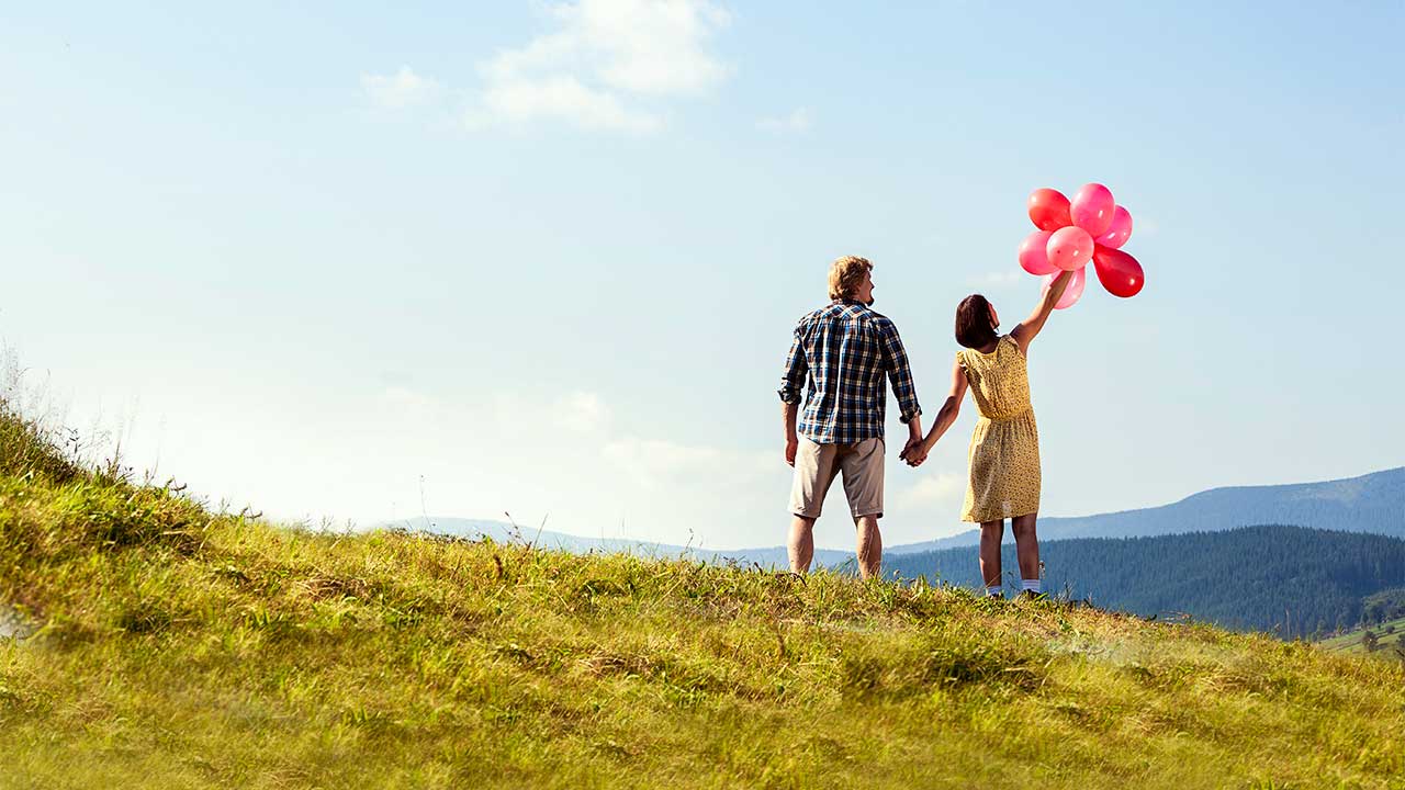 junge Paar steht auf einer Bergwiese, sie hält rote Ballone in der Hand