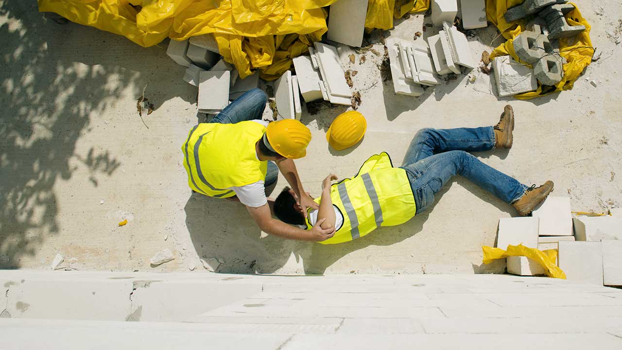 Zwei Bauarbeiter: Einer liegt verletzt am Boden, der andere hilft ihm.