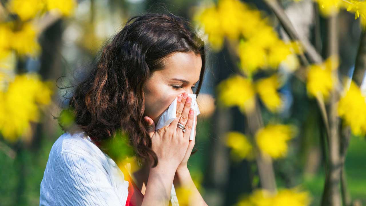 Frau niest in ein Taschentuch mit gelben Blumen im Hintergrund