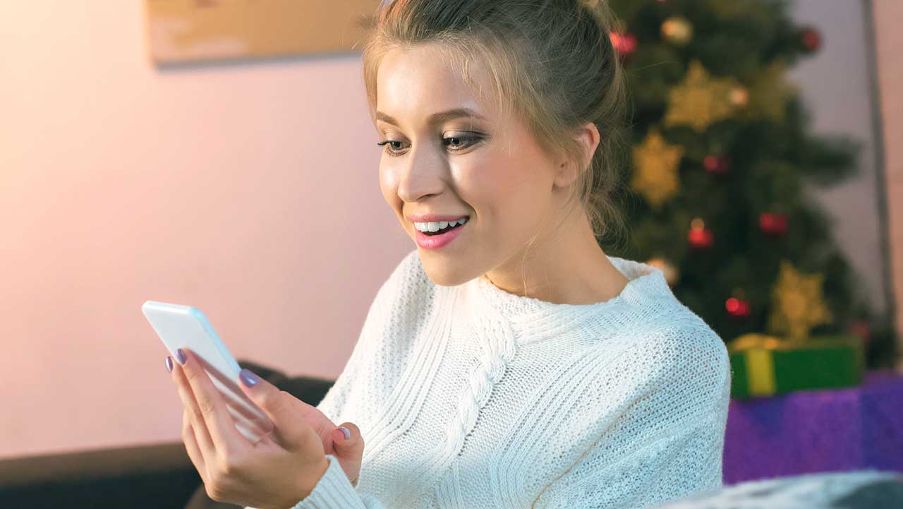 Frau sitzt auf Sofa mit Weihnachtsbaum im Hintergrund und hält Smartphone in der Hand