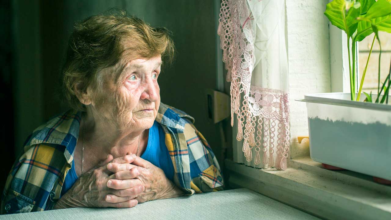 Ältere, einsame Frau sitzt an einem Tisch bei einem Fenster und sieht bedrückt aus