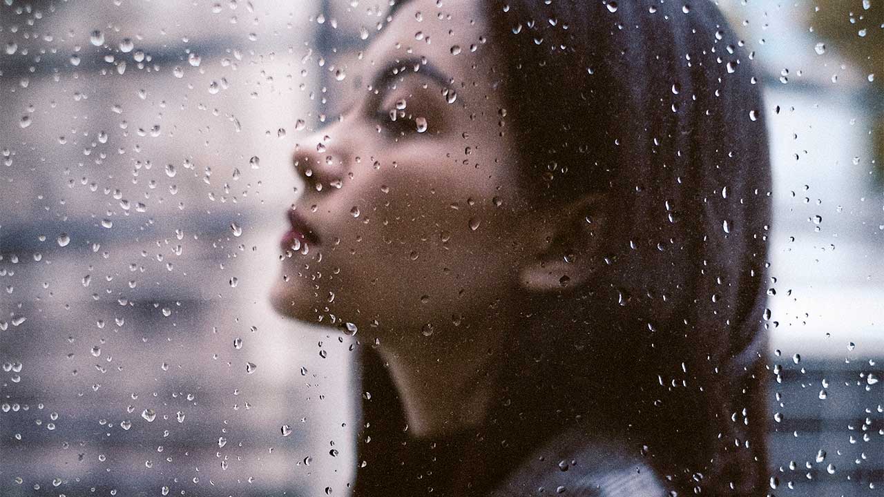 Ernste junge Frau hinter einer Glasscheibe mit Regentropfen