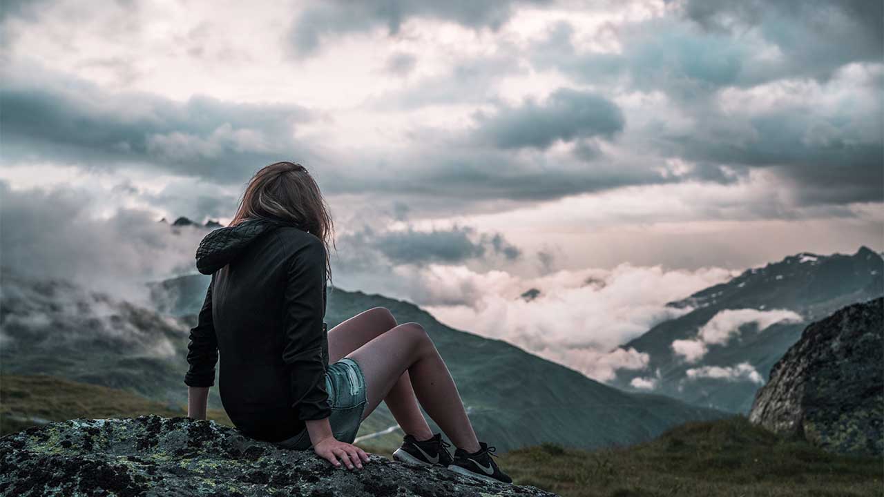 junge Frau sitzt in einer gebirgigen Landschaft und blickt hoch zum bewölkten HImmel