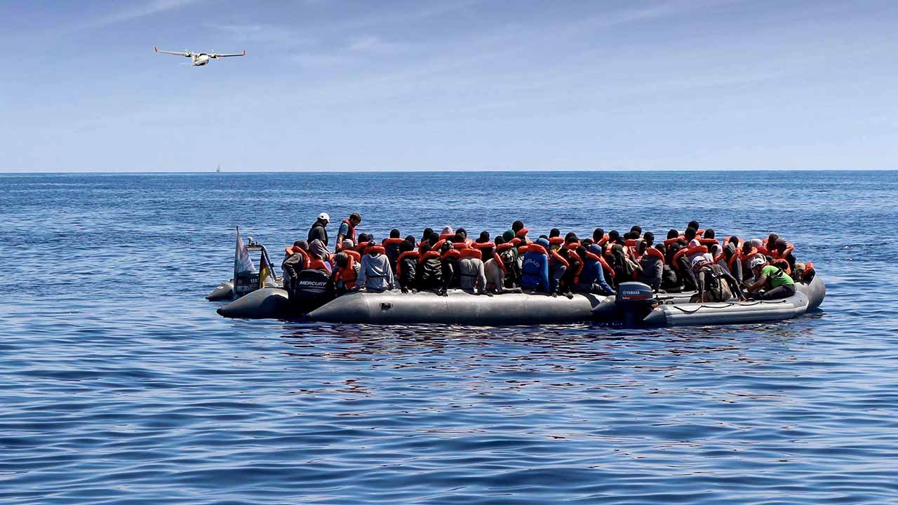 Flüchtlinge in einem Boot, im Hintergrund ein Erkundungsflugzeug