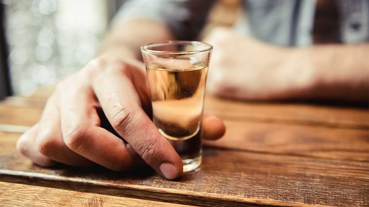männliche Hand auf einem Holztisch hält ein Gläschen Alkohol
