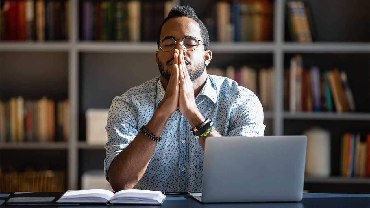 Mann beten in einem Büro, vor ihm ein Laptop und ein offenes Buch