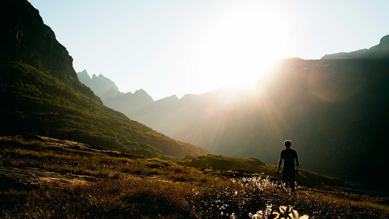 Mann auf den Lofoten, Norwegen wandert auf einer Wiese und blickt auf die Sonne, welche sich über einem Berggrat zeigt