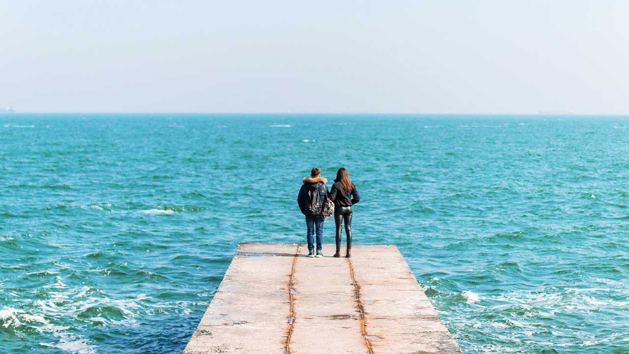 Rückansicht auf ein junges Paar, welches am Rand eines Piers mit Blick auf das Meer steht