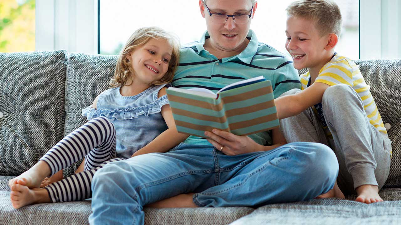 Vater sitzt mit Tochter und Sohn auf dem Sofa und liest aus einem Buch vor